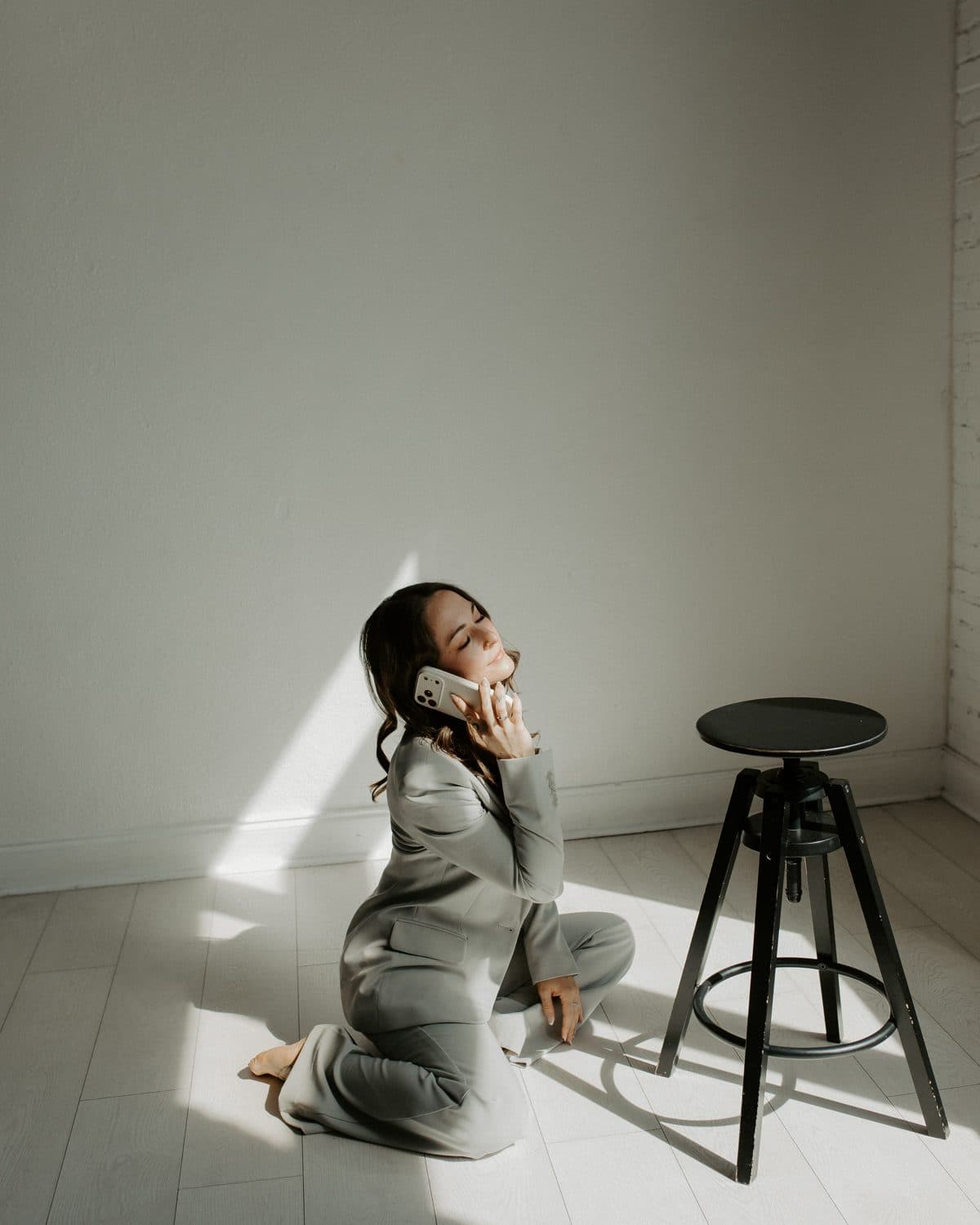 Woman in a grey suit kneeling on the floor on her phone beside a stool
